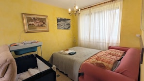 Ein Zimmer mit einem Bett, einem kleinen Tisch mit Stuhl, einem Babybett und zwei Bildern an der Wand.