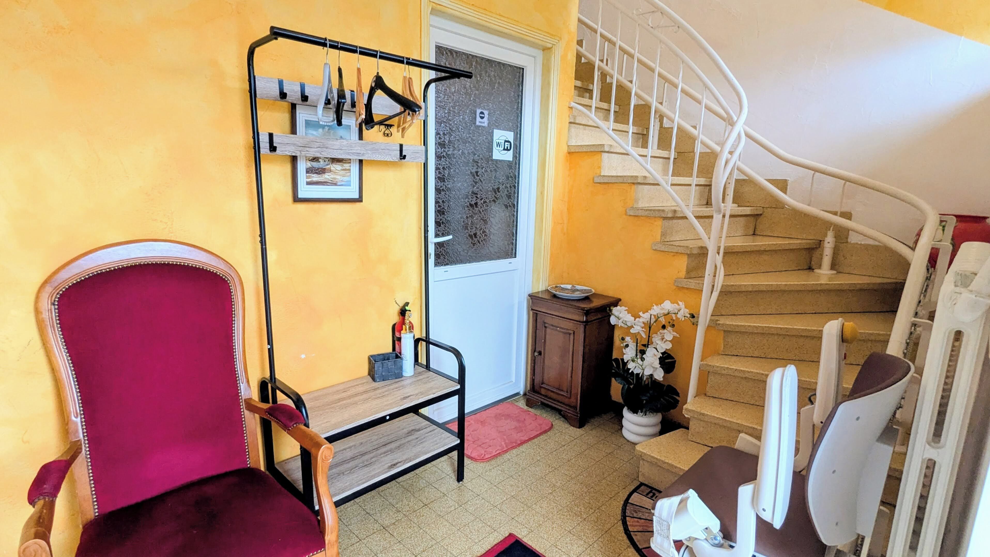 A room with a red chair, a wooden table, a metal rack with toiletries, a staircase, and a door with a 'WiFi' sign.