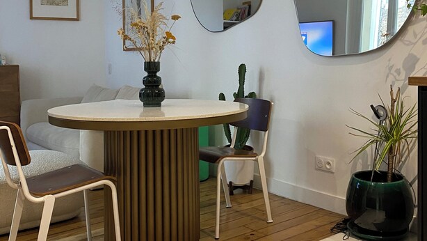 A dining area with a round table, two chairs, a vase with dried flowers, a cactus, and two mirrors on the wall.