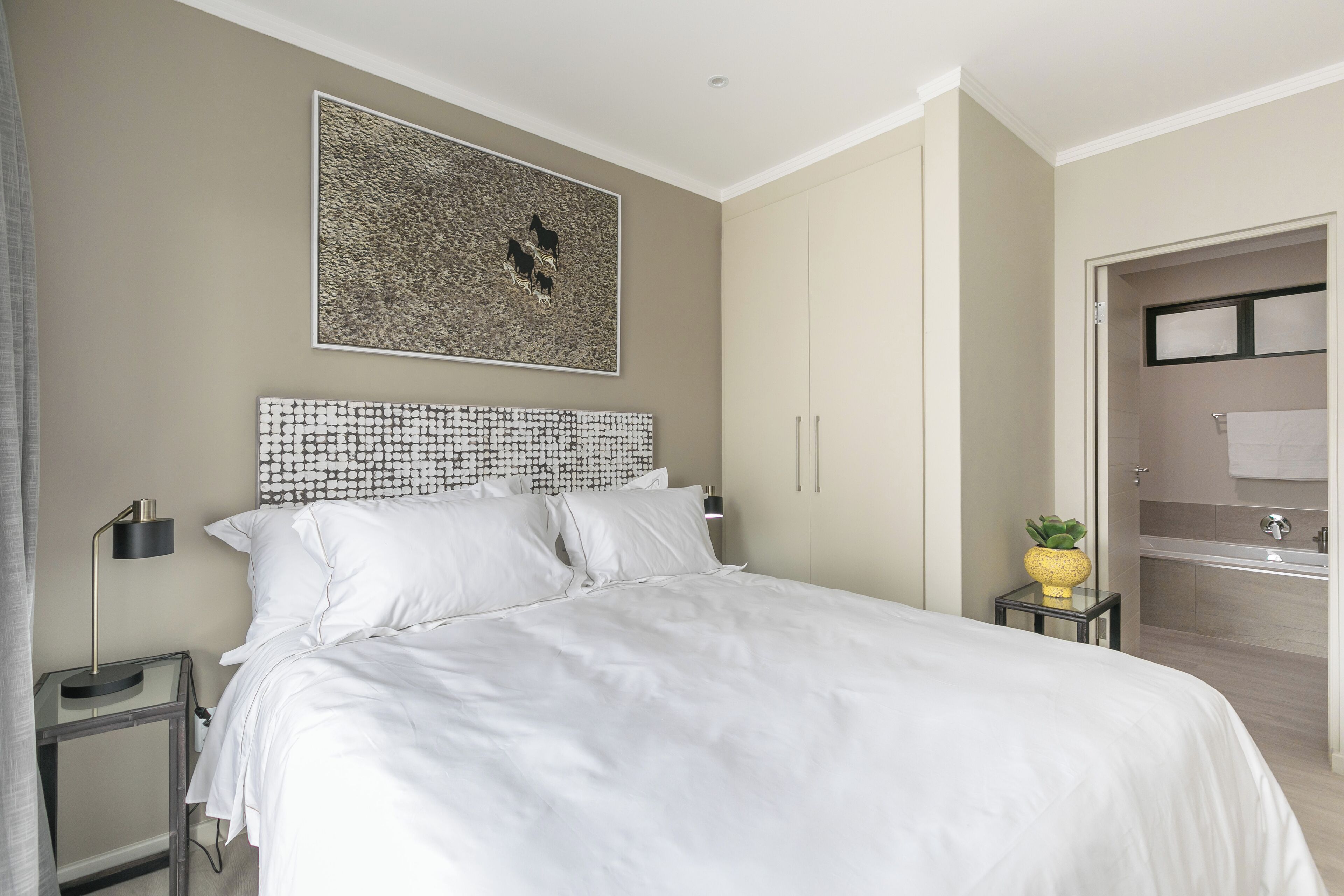 Un dormitorio moderno con una cama grande, un cabecero con diseño geométrico, una mesita de noche con lámpara y un baño con bañera y toallero.
