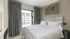 Ein modernes Hotelzimmer mit einem großen Bett, grauen Vorhängen, Blick auf ein Gebäude durch eine Glastür und einem gerahmten abstrakten Kunstwerk an der Wand.