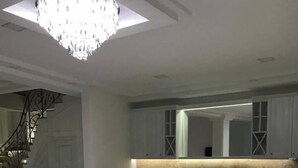 A spacious living room with a large chandelier, a comfortable armchair, and a kitchen area with white cabinetry and a wooden countertop. - Adams Hotel (Yerevan)