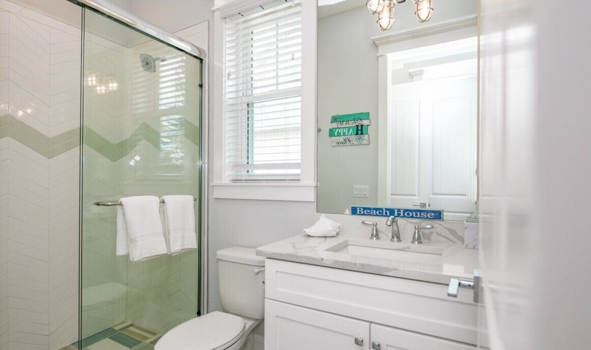 Een badkamer met een douchecabine van glas, een toilet, een wastafel met spiegel en een raam met zonwering.