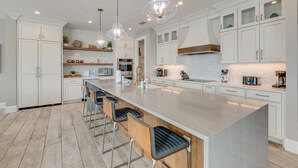 A modern kitchen with white cabinets, a large island with bar stools, pendant lighting, and stainless steel appliances.