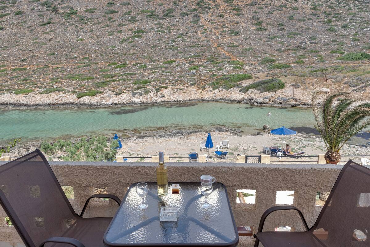 A table with two chairs, a bottle, and two glasses on a balcony overlooking a beach with a rocky landscape and a clear turquoise sea.