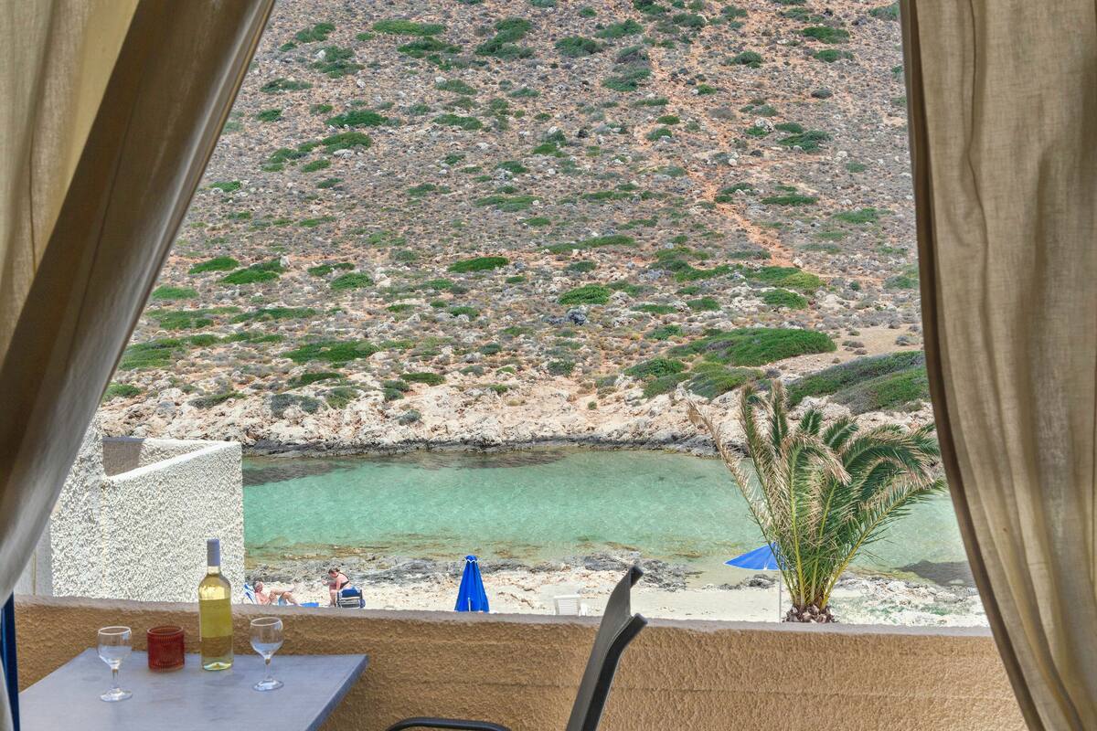 A table with a bottle and glasses, a blue umbrella, and a view of a rocky beach and clear water.
