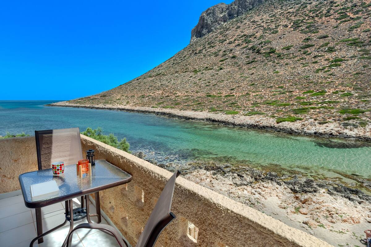 A balcony with a table and chairs overlooking a clear blue sea and a rocky hillside.