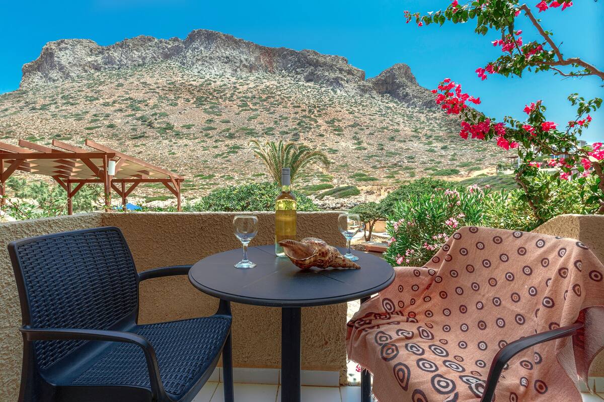 A round table with two chairs, a bottle, and two glasses on a balcony with a view of a mountainous landscape.