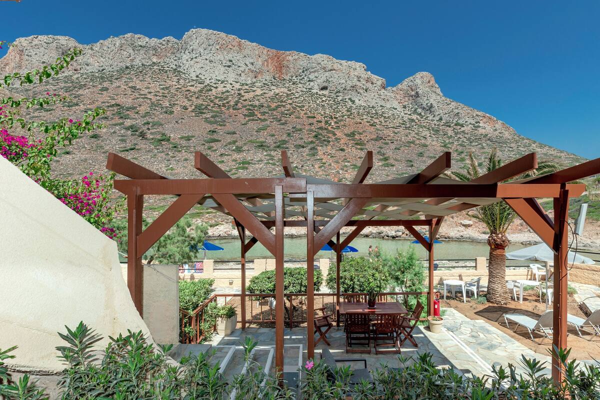 A covered outdoor seating area with tables and chairs, a palm tree, and a mountainous backdrop.