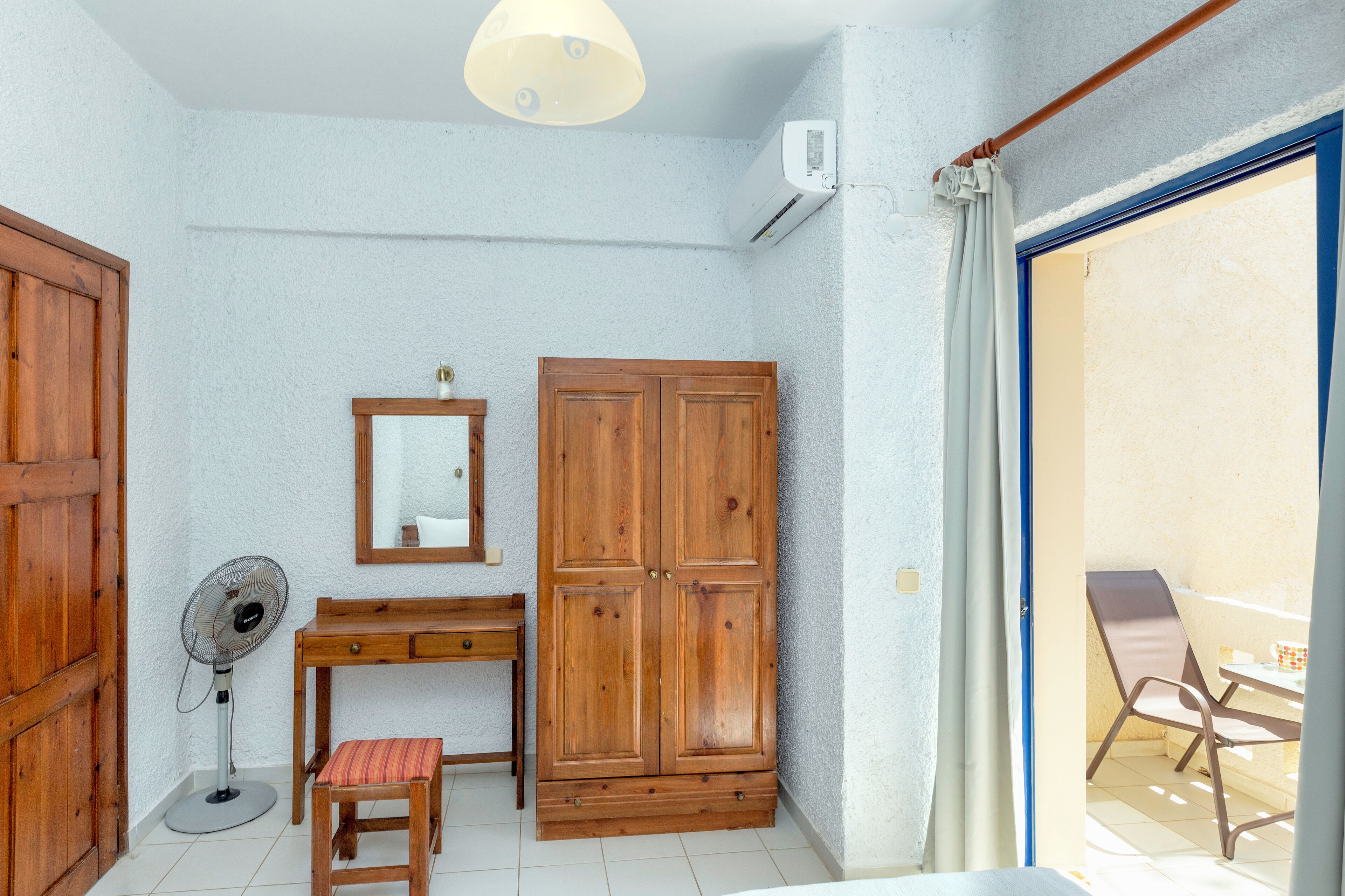 A room with a wooden wardrobe, a desk with a chair, a standing fan, and a window with a curtain.