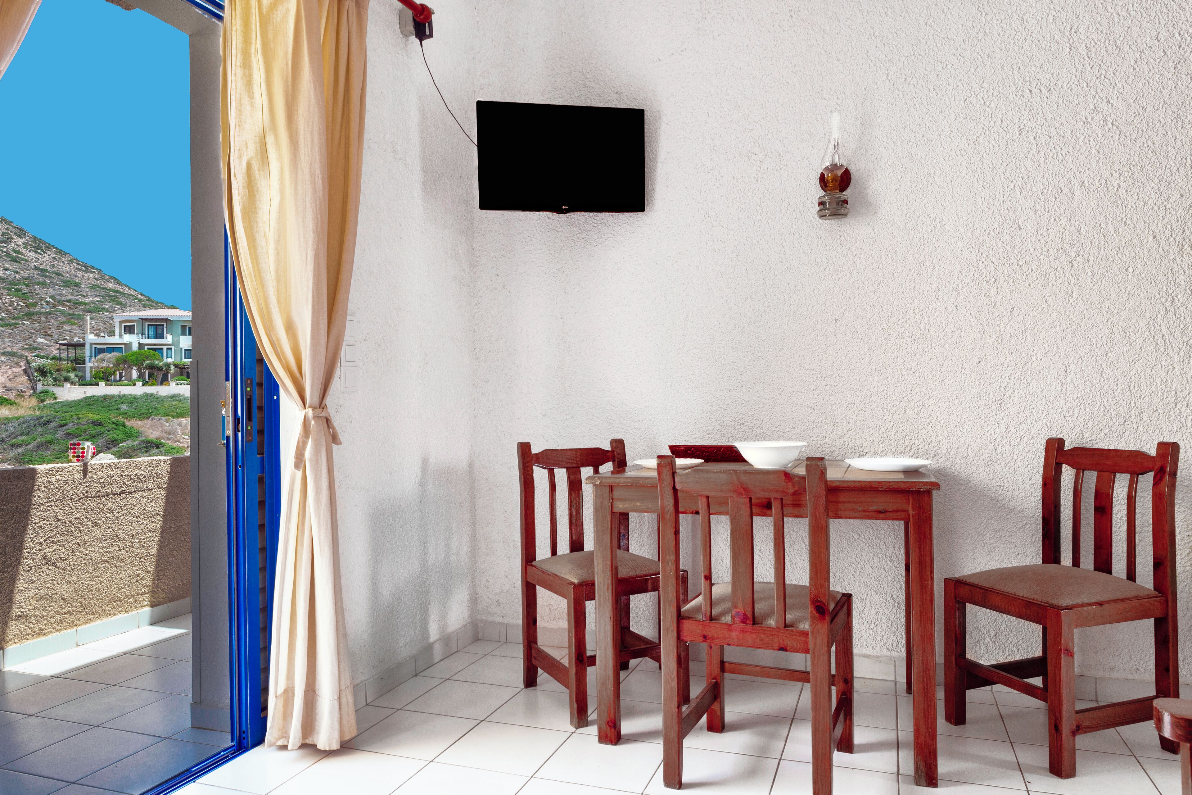 A dining area with a table and chairs, a TV mounted on the wall, and a view of a hillside.