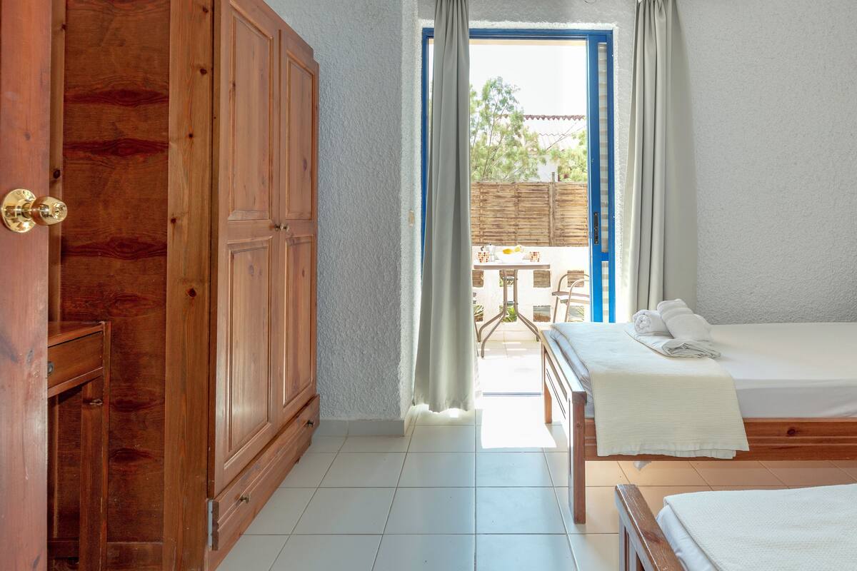A bedroom with a wooden wardrobe, a bed with a white bedspread, and a view of an outdoor patio with a table and chairs.