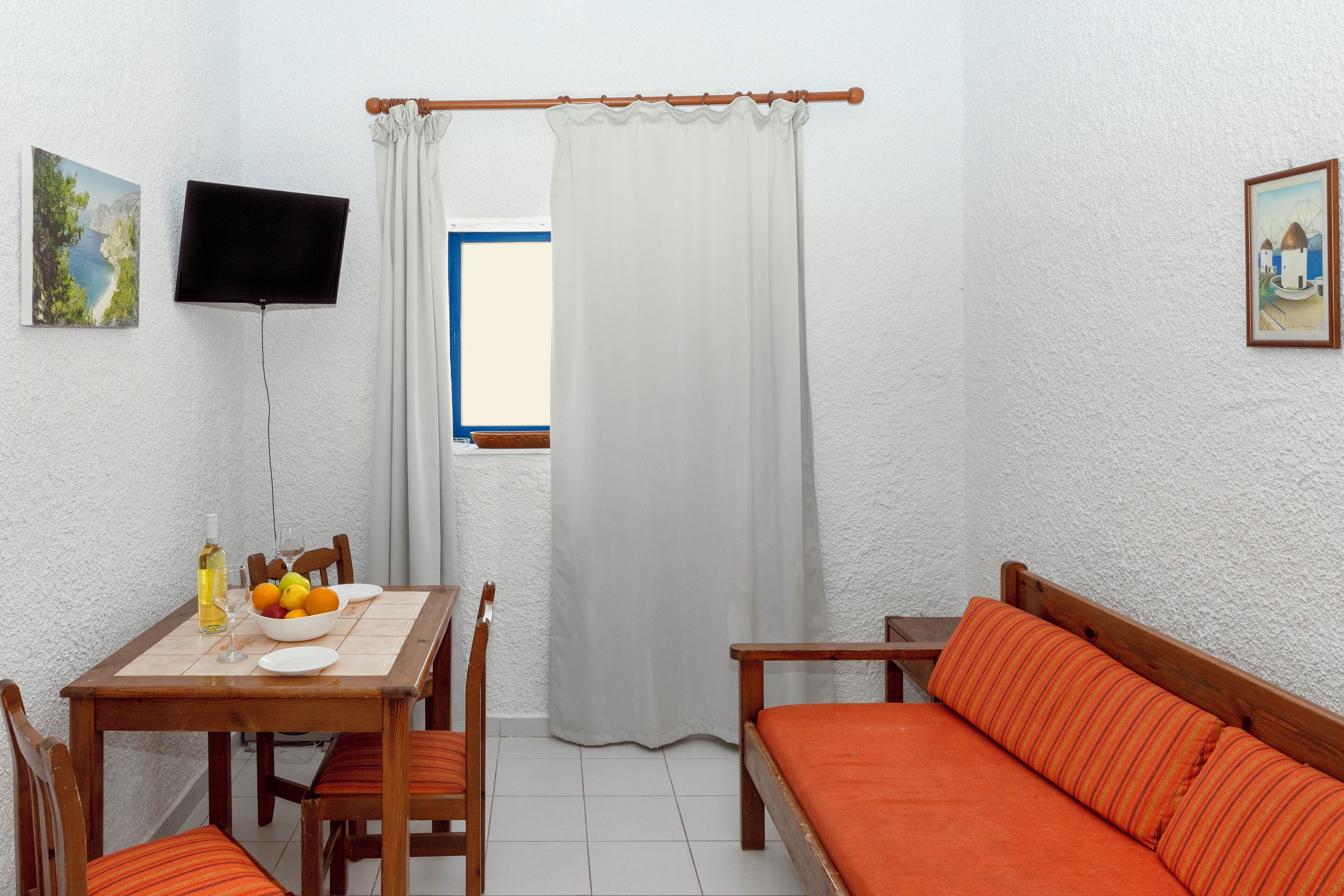 A room with a dining table, chairs, a sofa, a TV mounted on the wall, and a window with curtains.
