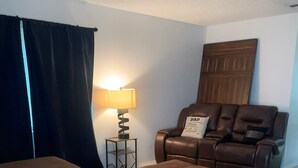 Ein Wohnzimmer mit braunem Ledersofa, passendem Hocker, einem kleinen Beistelltisch mit Lampe und einem dunklen Vorhang.