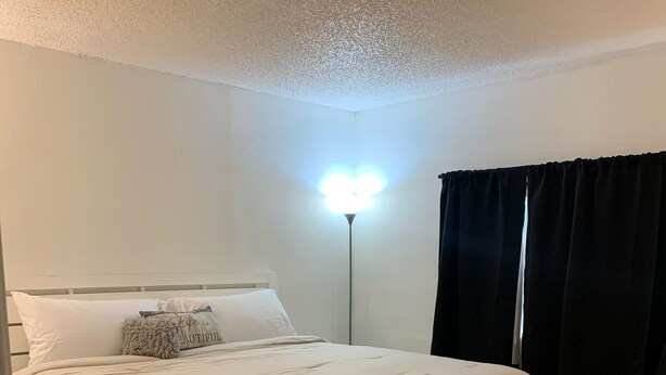 Ein ordentlich bezogenes Bett mit weißer Bettwäsche und einem flauschigen Kissen, ein Nachttisch, eine Stehlampe und ein Fenster mit dunklen Vorhängen.