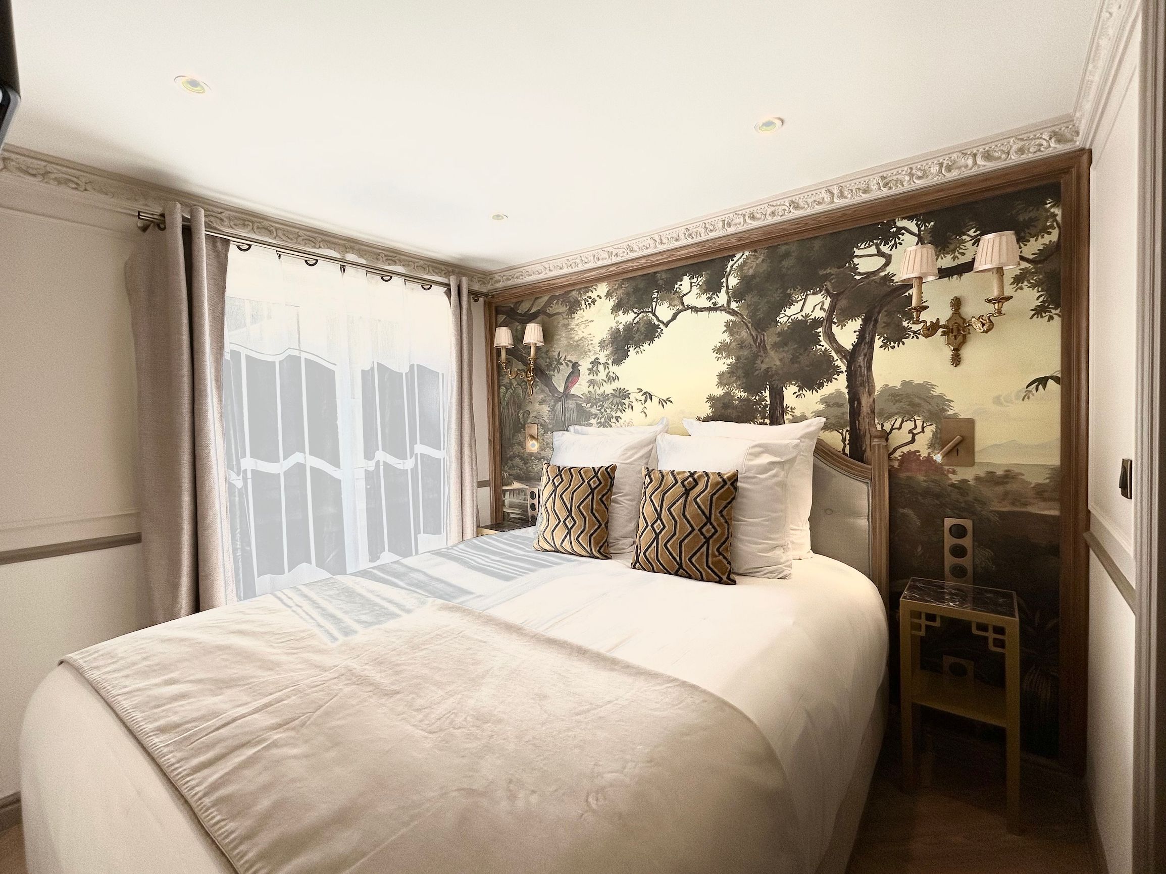 Ein Hotelzimmer mit einem großen Bett, Blick auf Bäume durch ein Fenster und einem kleinen Tisch mit einer Lampe.