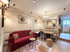 A spacious living area with a red sofa, a wooden coffee table, and a dining table with chairs. There are chandeliers and wall-mounted lamps providing lighting. - Merveil - Signature Suite - Tour Eiffel - Bourgogne (Paris)