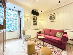 A living room with a red sofa, a small round table, a chair, and a large window with a blue leaf pattern.