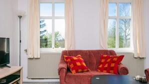 A cozy living room with a red sofa, two colorful pillows, a flat-screen TV on a wooden stand, and a large window with curtains. - 6 Person Holiday Home in Skaerbaek (Skaerbaek)