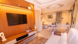 A modern living room with a flat-screen TV, wooden panel wall, white sofa, dining area with chairs, and a patterned rug. - Apto no Coração do Capivari e Perto de Tudo! (Campos do Jordão)