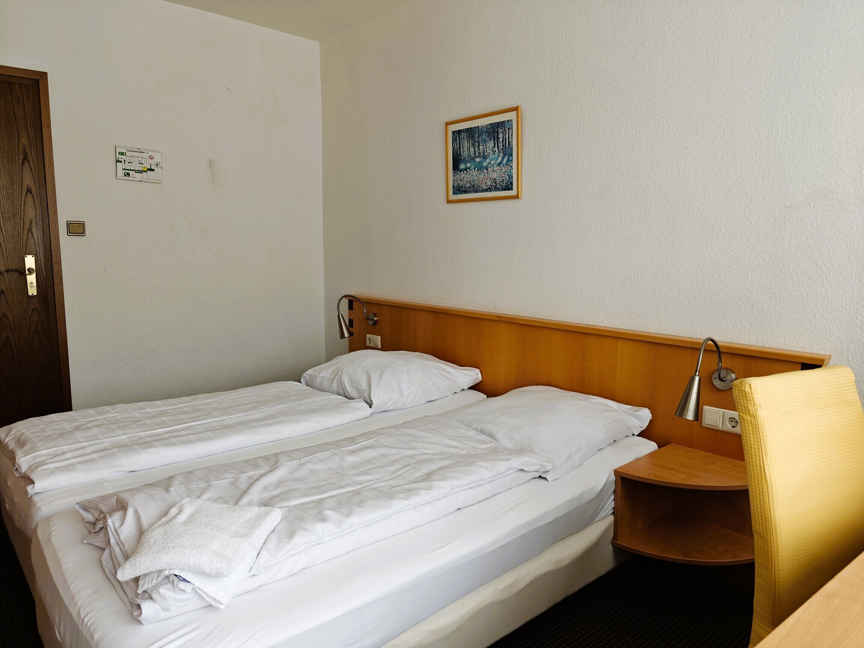 Cama doble con ropa de cama blanca, cabecera de madera, mesita de noche con lámpara y un cuadro en la pared.