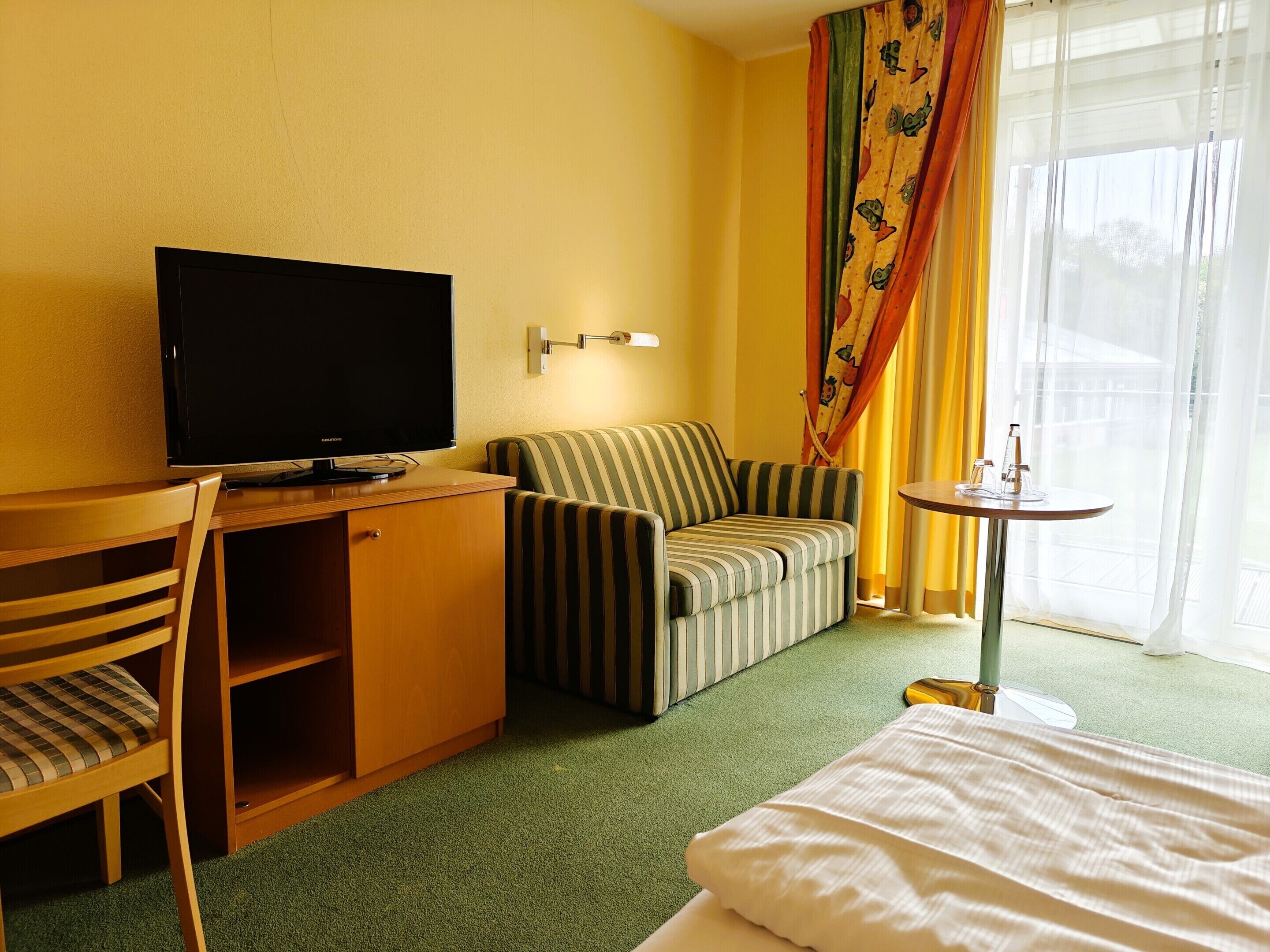 A hotel room with a television on a wooden desk, a striped sofa, a small round table with a lamp, and a bed with white bedding.