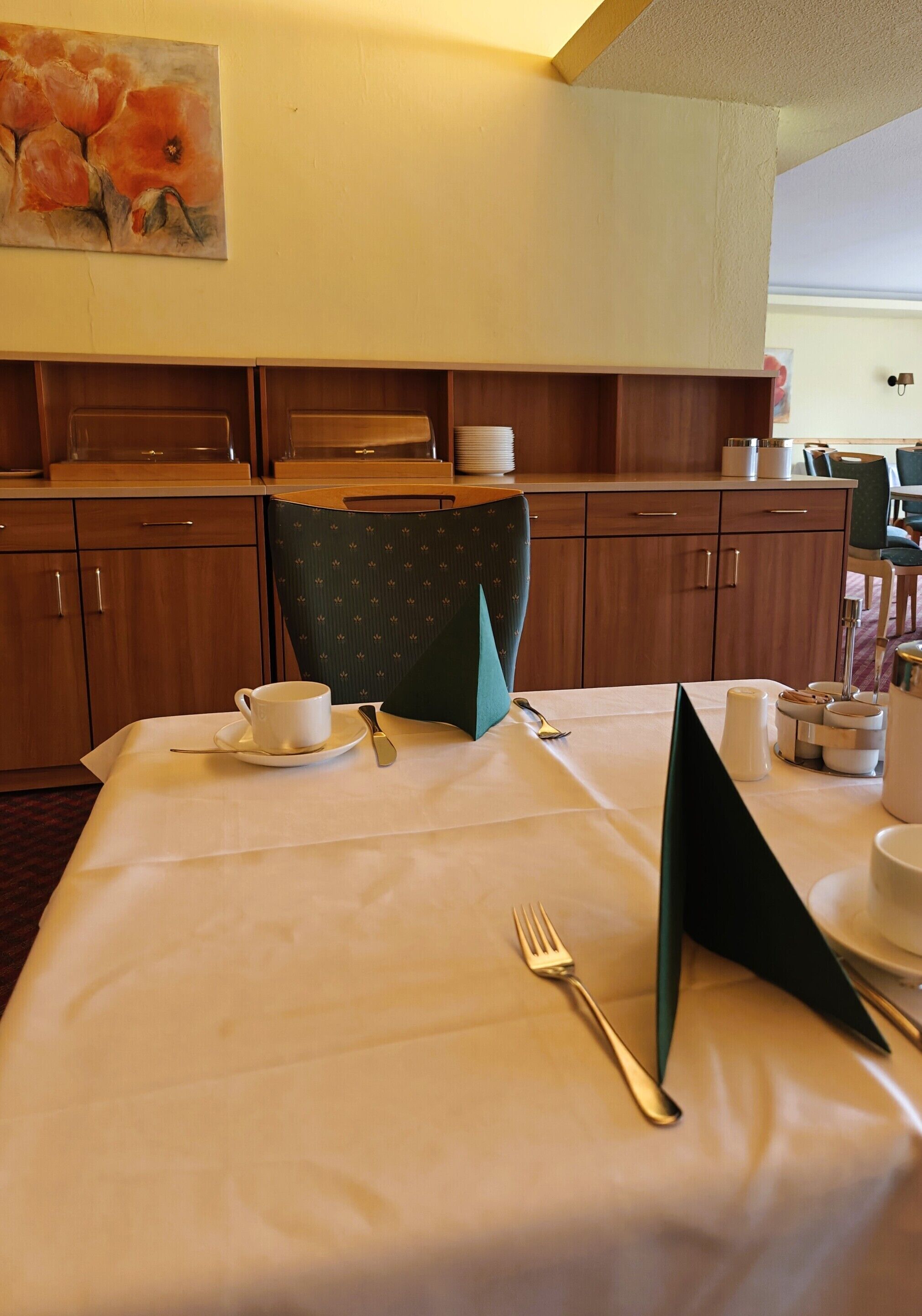 A neatly set dining table with a cup, saucer, and cutlery, a green chair, and a painting of flowers on the wall.