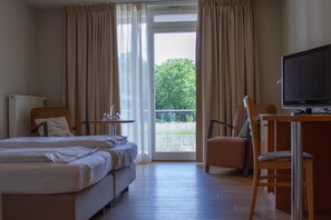 Habitación de hotel con cama, dos sillas, escritorio con televisor y balcón con cortinas.