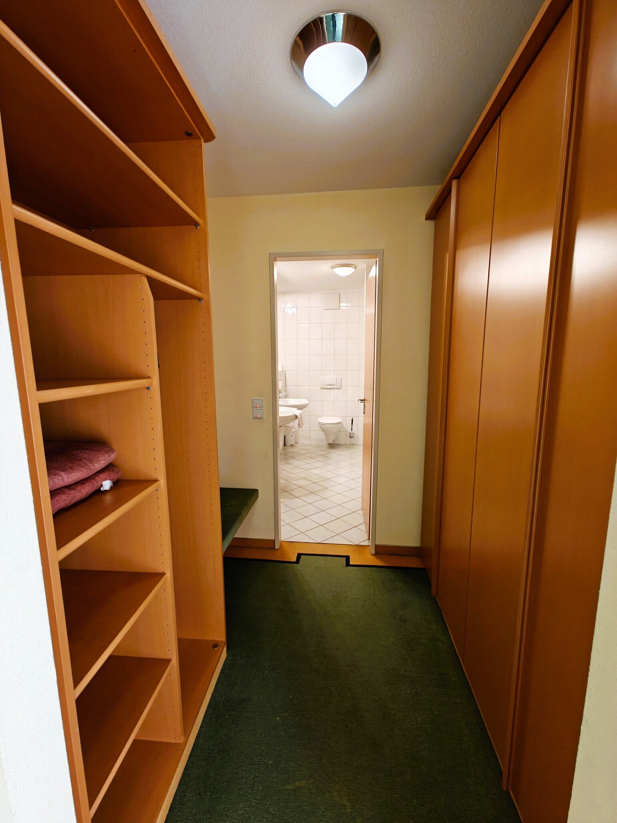 Un couloir étroit, avec des étagères en bois d’un côté et une porte donnant accès à une salle de bain.