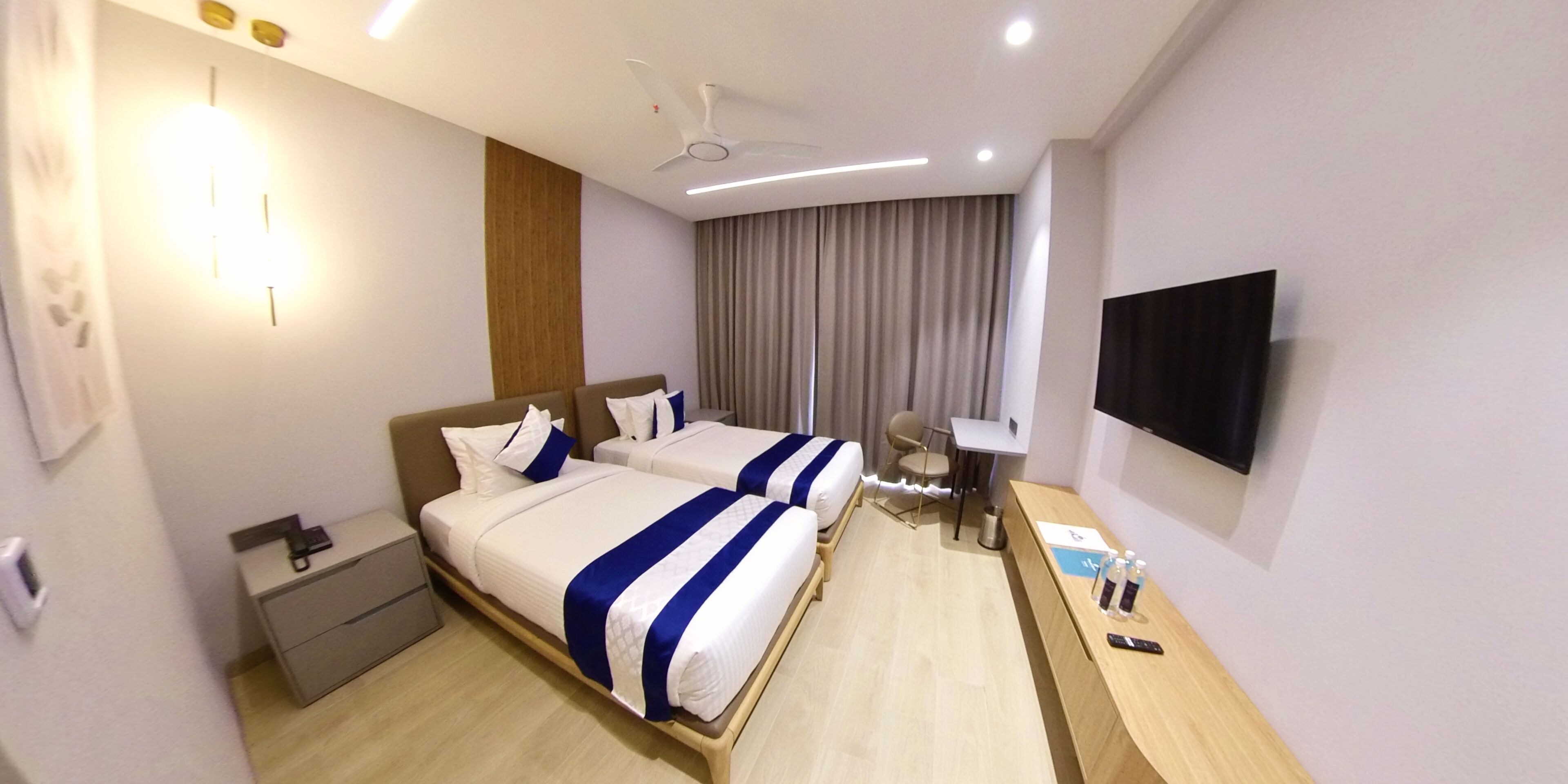 A hotel room with two beds, a desk, a chair, a TV, and a ceiling fan.