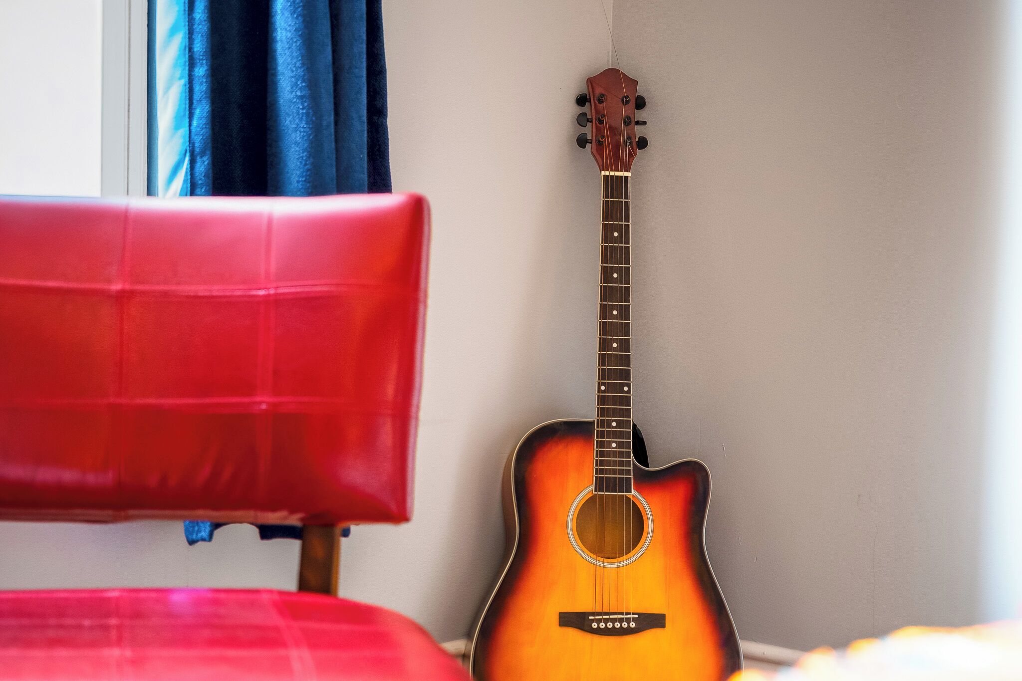 A guitar leaning against a wall in a room with a red chair.