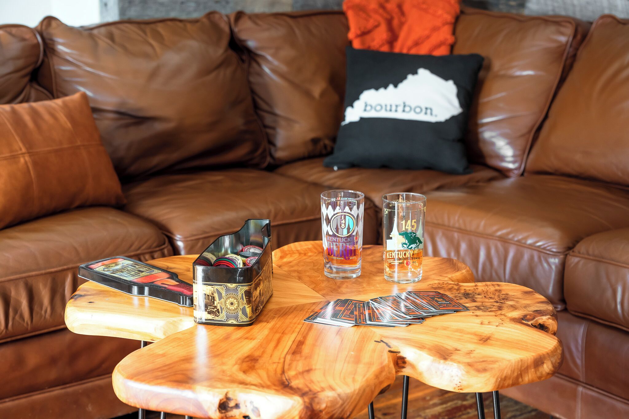 A brown leather couch with a black pillow labeled "bourbon" and two glasses on a wooden table with playing cards and a cigar box.