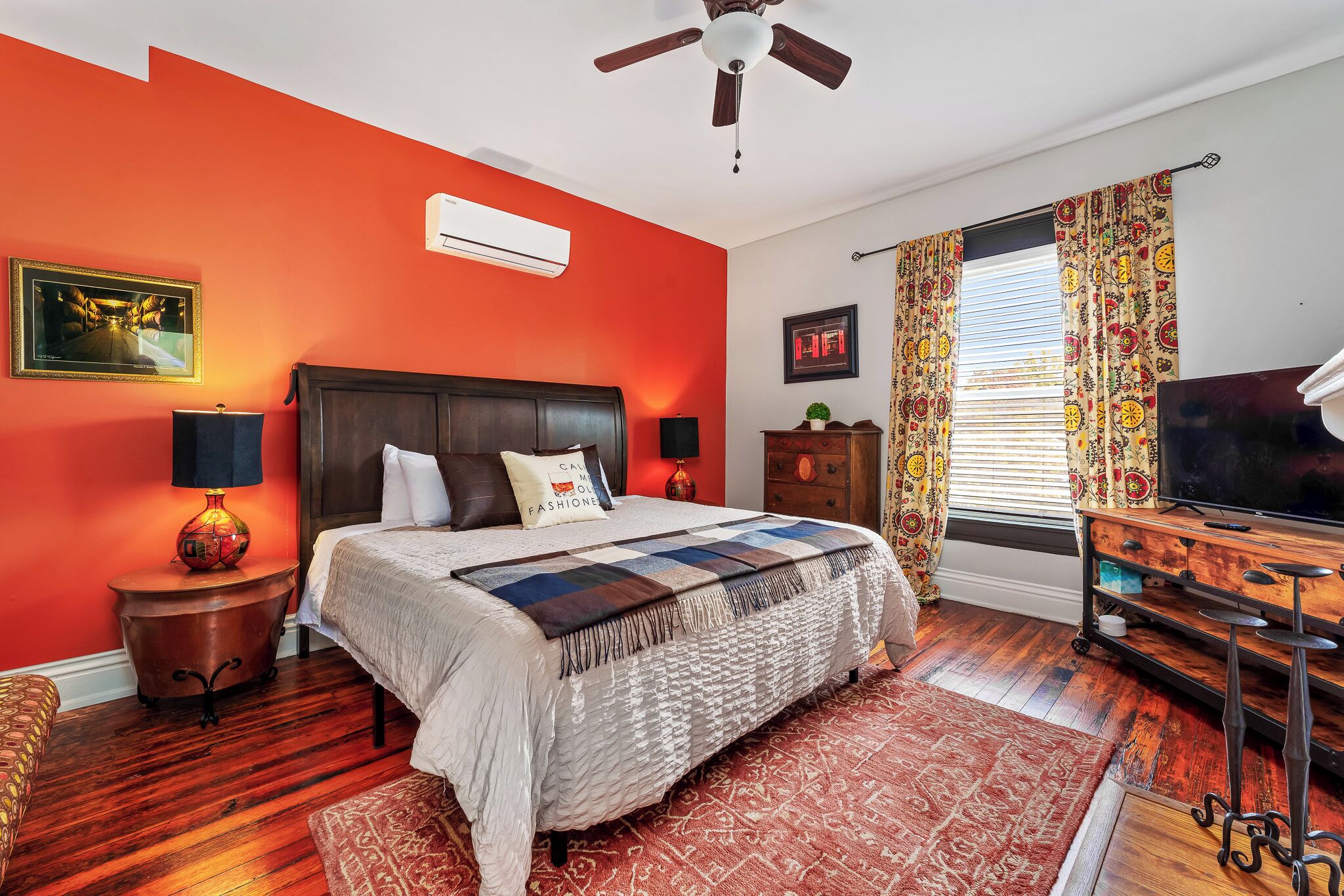 A bedroom with a large bed, a ceiling fan, a TV, and a window with curtains.