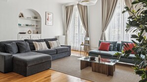 A modern living room with a sectional sofa, a glass coffee table, and a side table with a lamp. There are two windows with curtains, a white console table with decorative items, and a plant in the corner. - Magnificent apartment II - 1BR/4P - Opéra-Bourse (Paris)