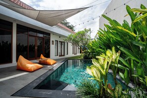 A modern outdoor pool area with a clear pool, orange bean bags, and lush greenery.