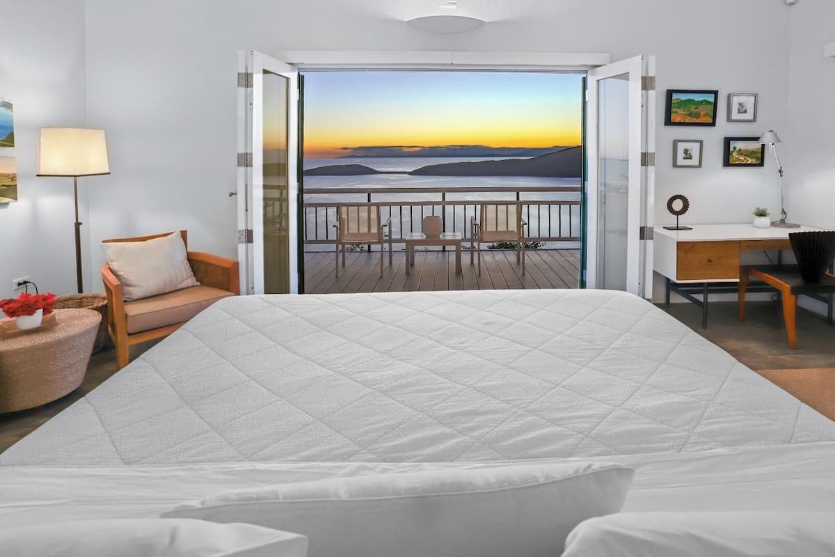 Un dormitorio con cama, escritorio, sillón, sofá y balcón con vista al mar al atardecer.
