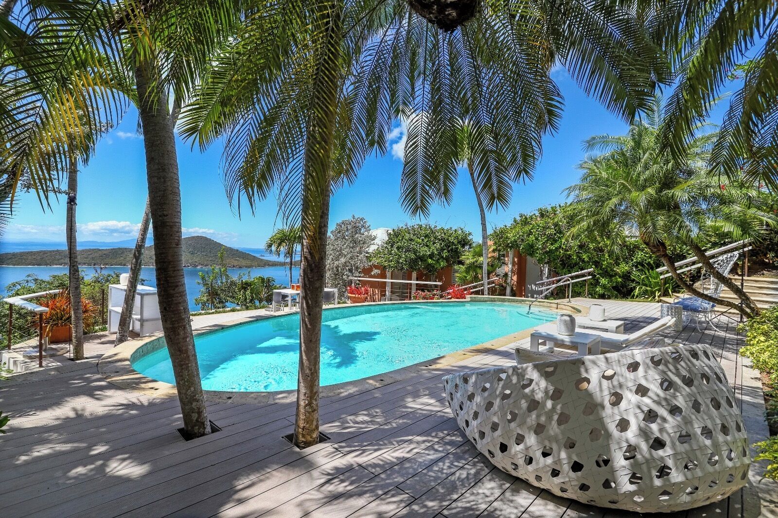 A pool area with a clear view of the sea and mountains, surrounded by palm trees and outdoor seating.