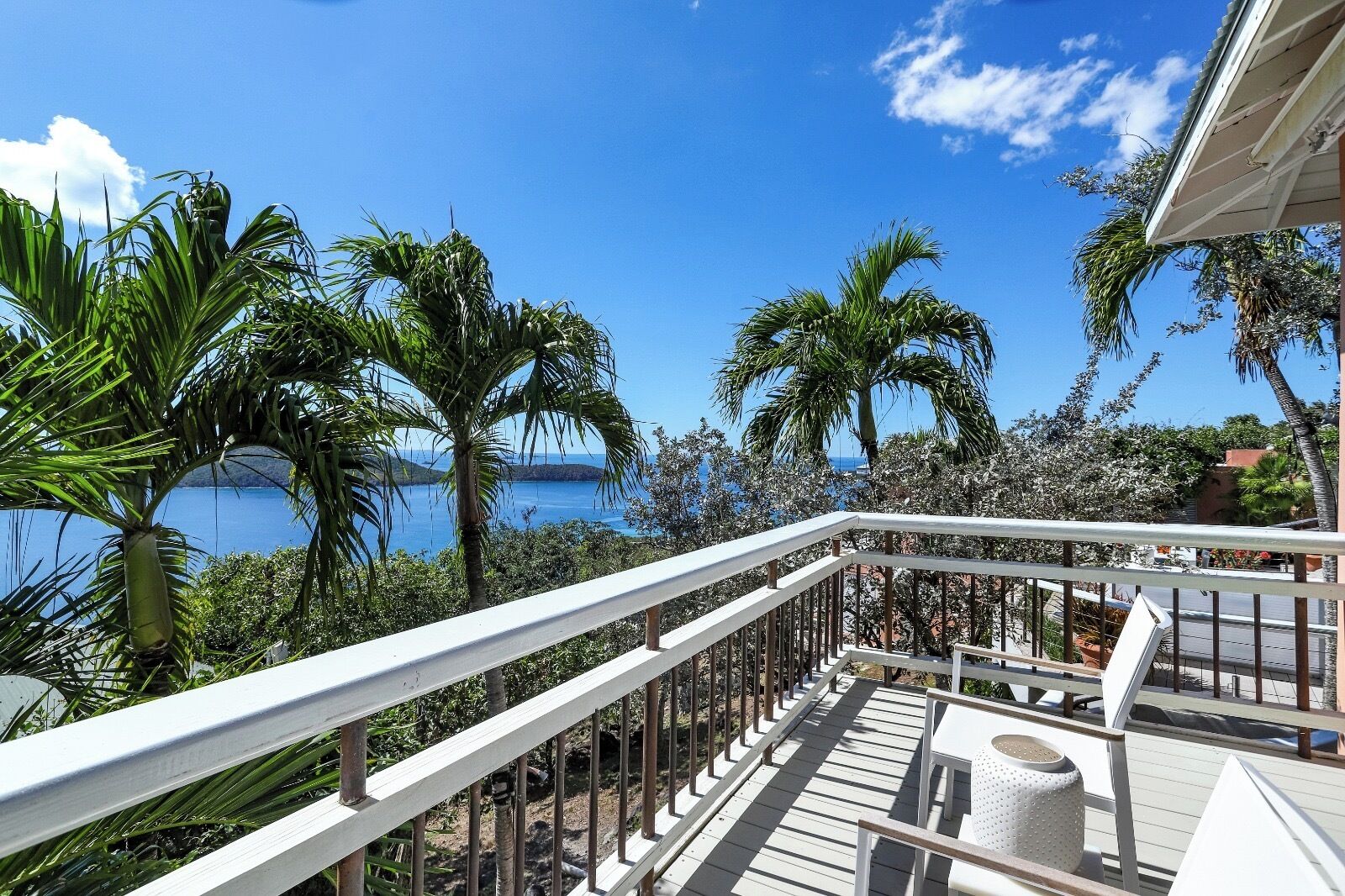 A balcony with a white railing overlooking a coastal view with palm trees and the sea.