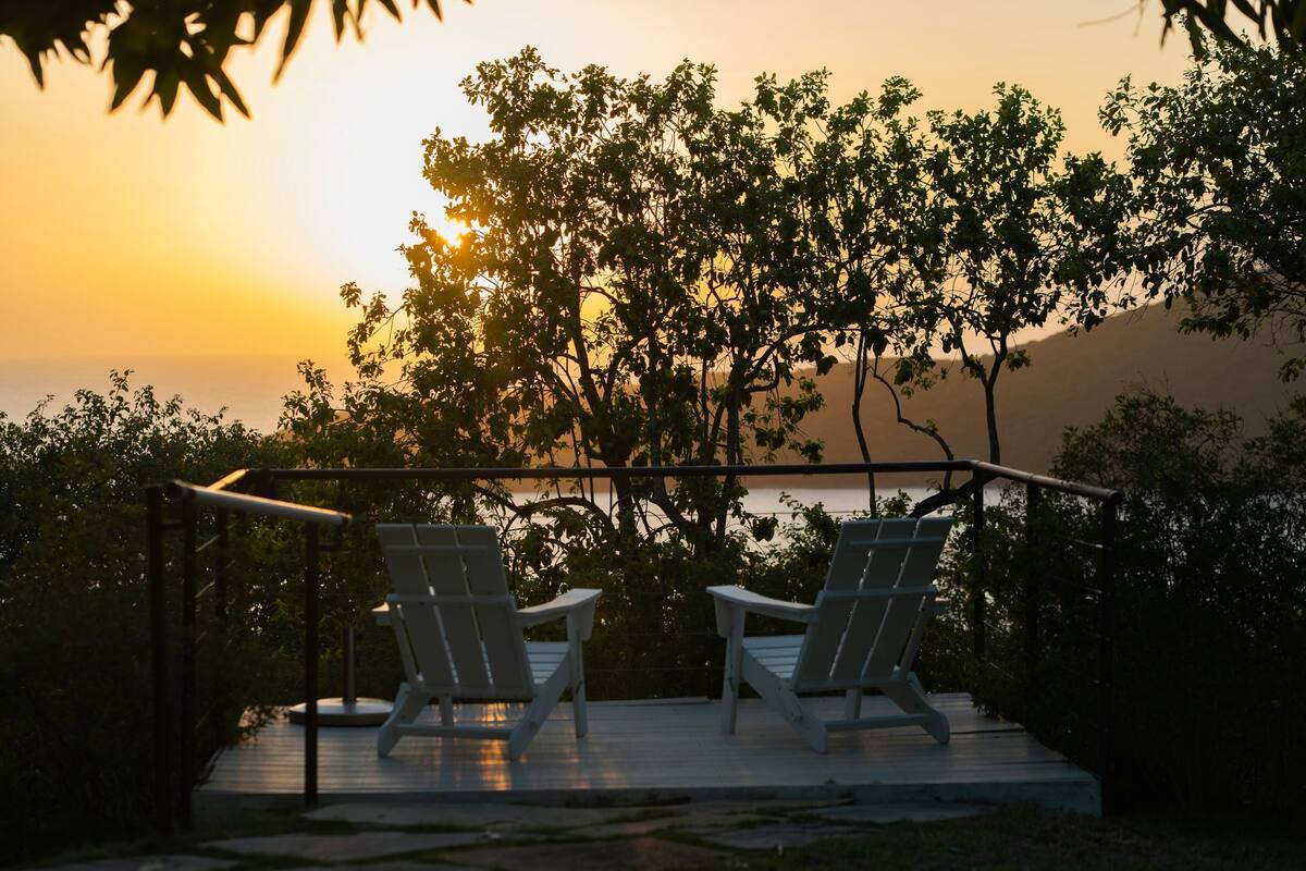 Dos sillas Adirondack blancas sobre una terraza de madera con barandilla metálica, con vistas a un atardecer y árboles.