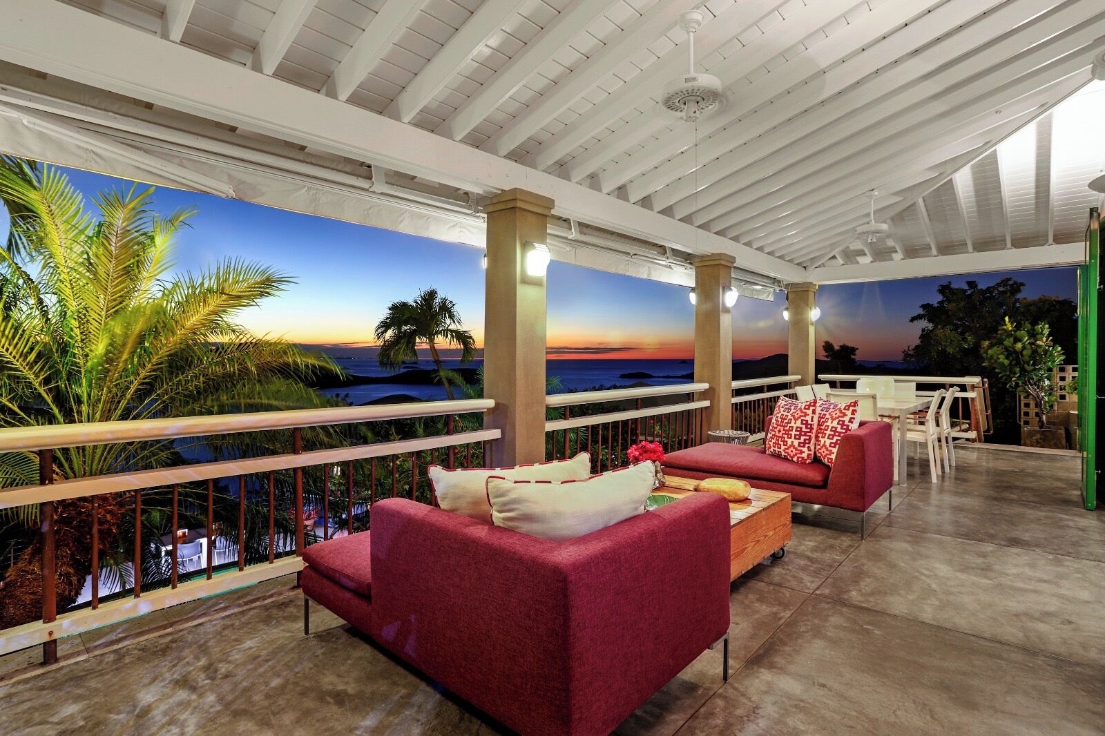 A covered outdoor seating area with red sofas and a view of the ocean at sunset.