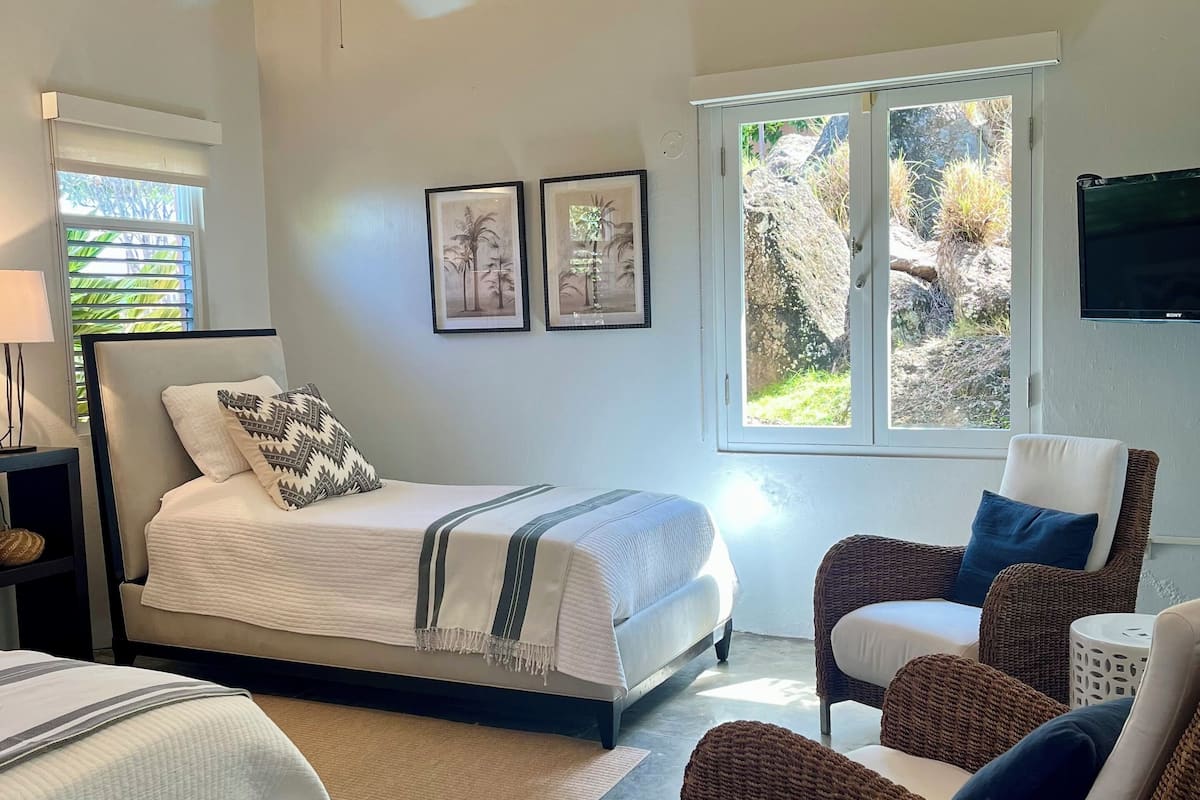 Un dormitorio con una cama, dos cuadros enmarcados, un ventilador de techo, una ventana con persianas y vistas al exterior.