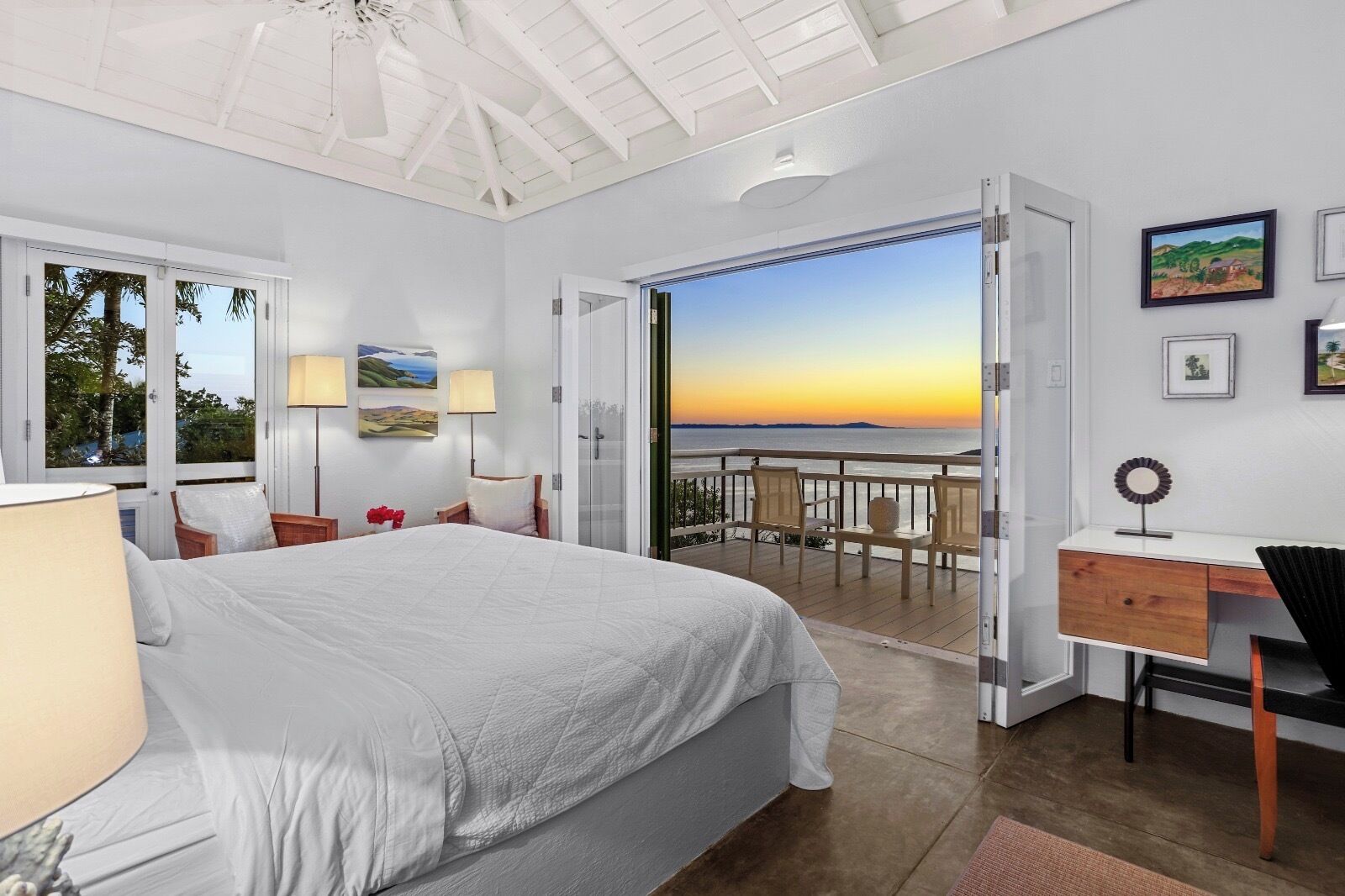 A bedroom with a large bed, a desk, and a balcony with a view of the sunset.