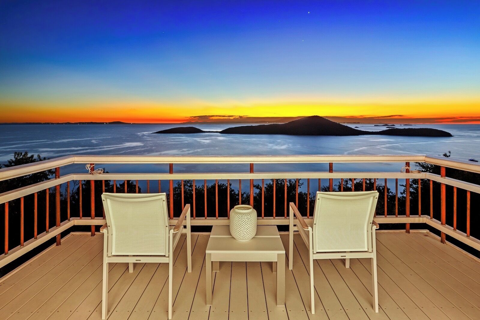 Un balcón con dos sillas y una mesa, con vistas a una puesta de sol sobre el agua y islas a lo lejos.