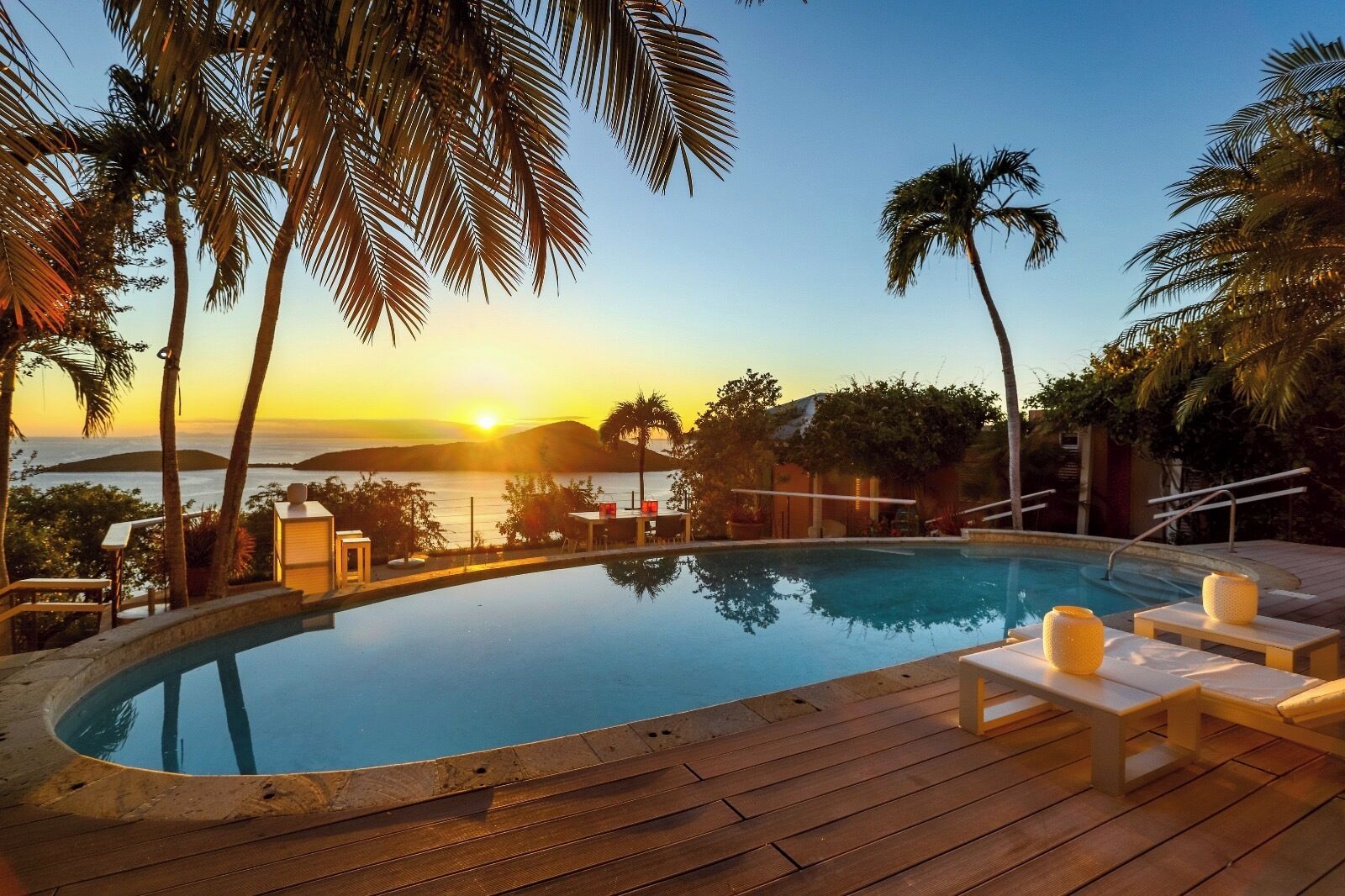 Una piscina con lettini e un pontile in legno, circondata da palme e con vista su un tramonto sull'oceano.
