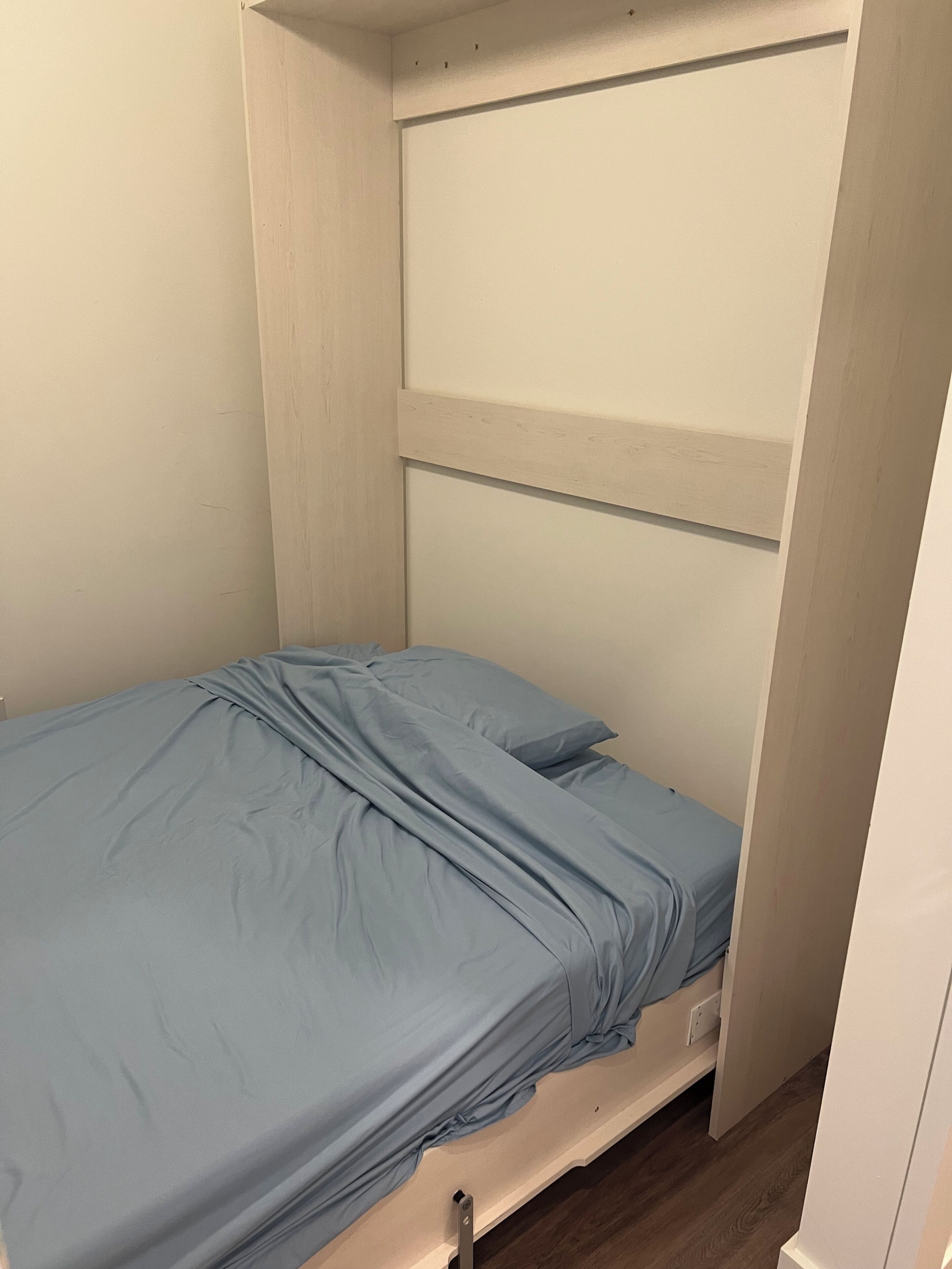 A small, single-bed room with a simple, light-colored headboard and a wooden floor.