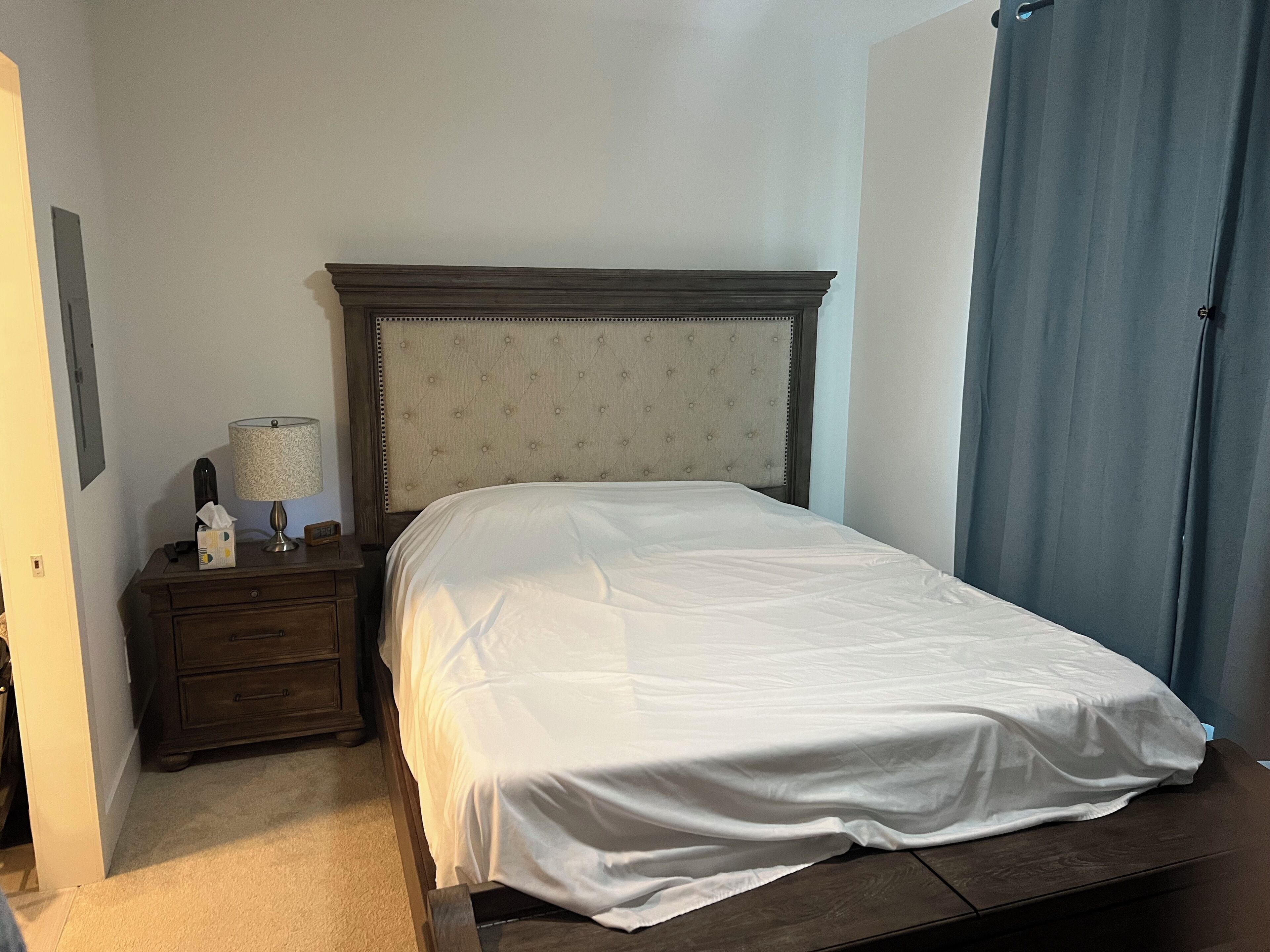 A neatly made bed with a headboard, a bedside table with a lamp, and a curtain on the window.