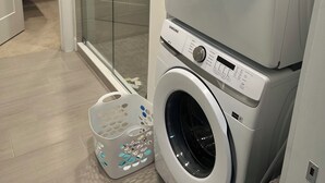 Un espace buanderie comprenant deux machines à laver superposées, une cabine de douche en verre et un panier à linge orné de motifs floraux.