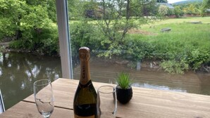 A wooden table with a bottle of CANTI wine and two glasses, overlooking a river and greenery.