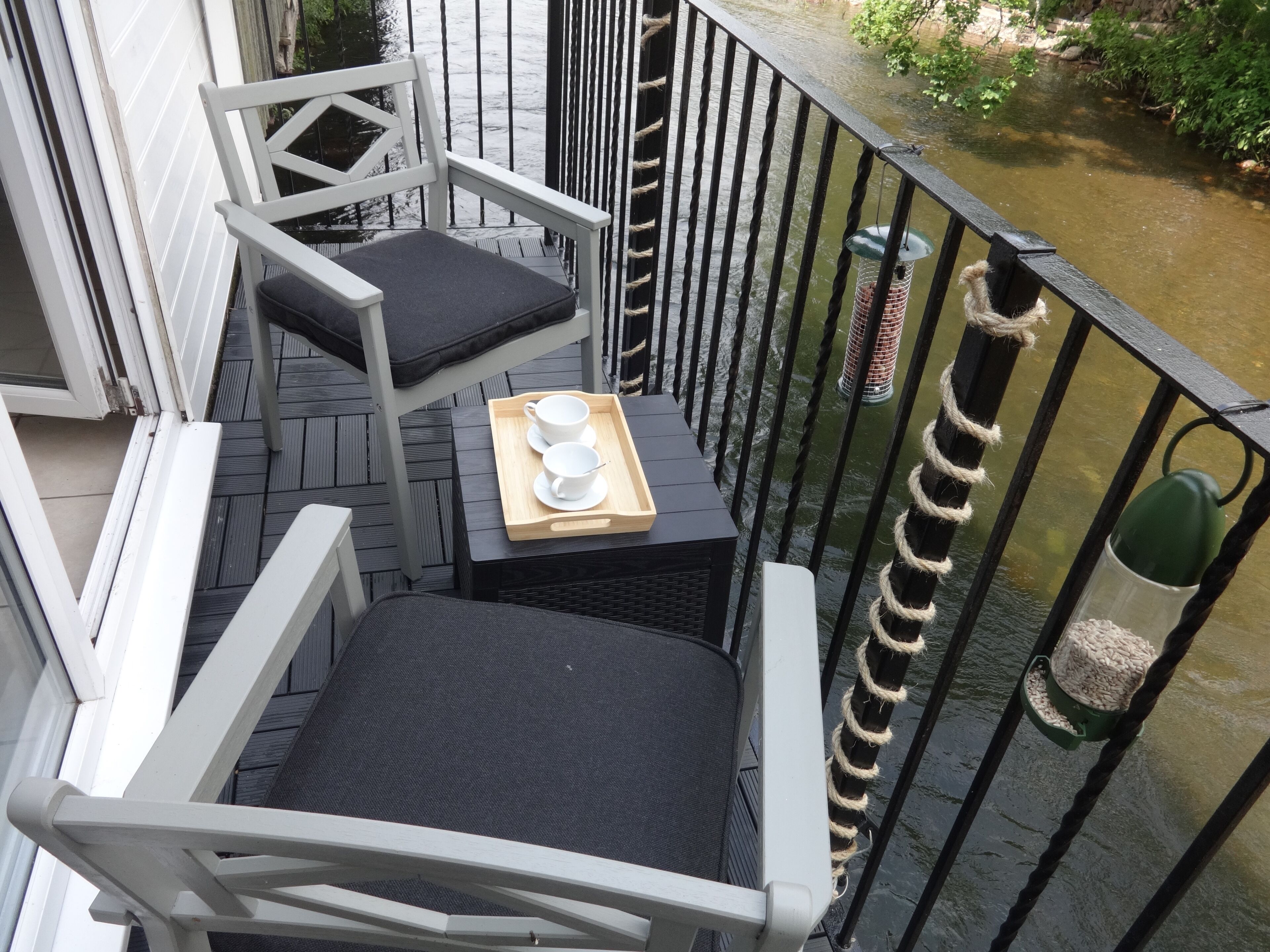 Balcón con mesa dispuesta, dos tazas, una bandeja, una silla y barandilla con vista a un elemento acuático.