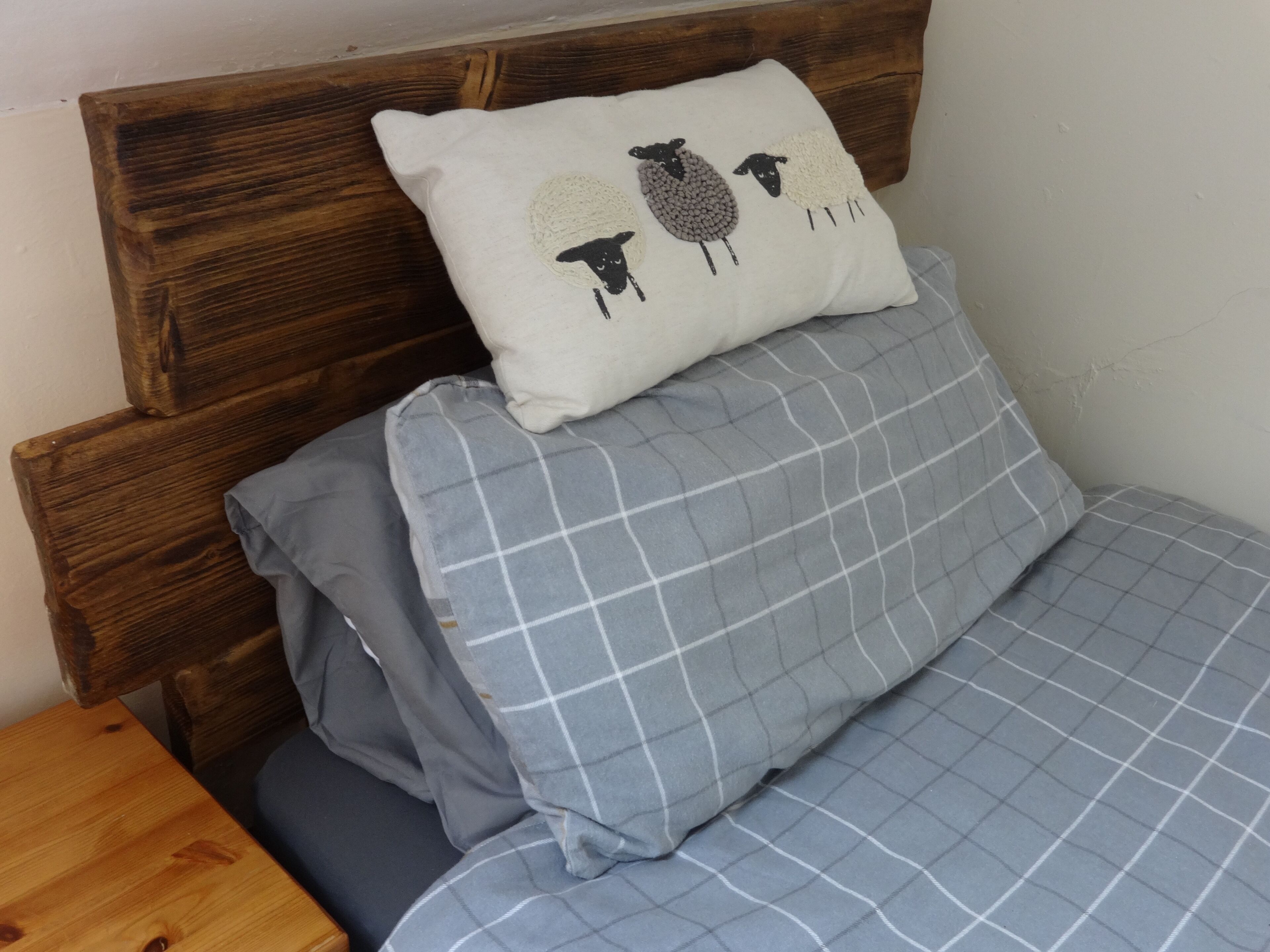 A bed with a wooden headboard, a pillow with a sheep design, and a blue checkered bedspread.